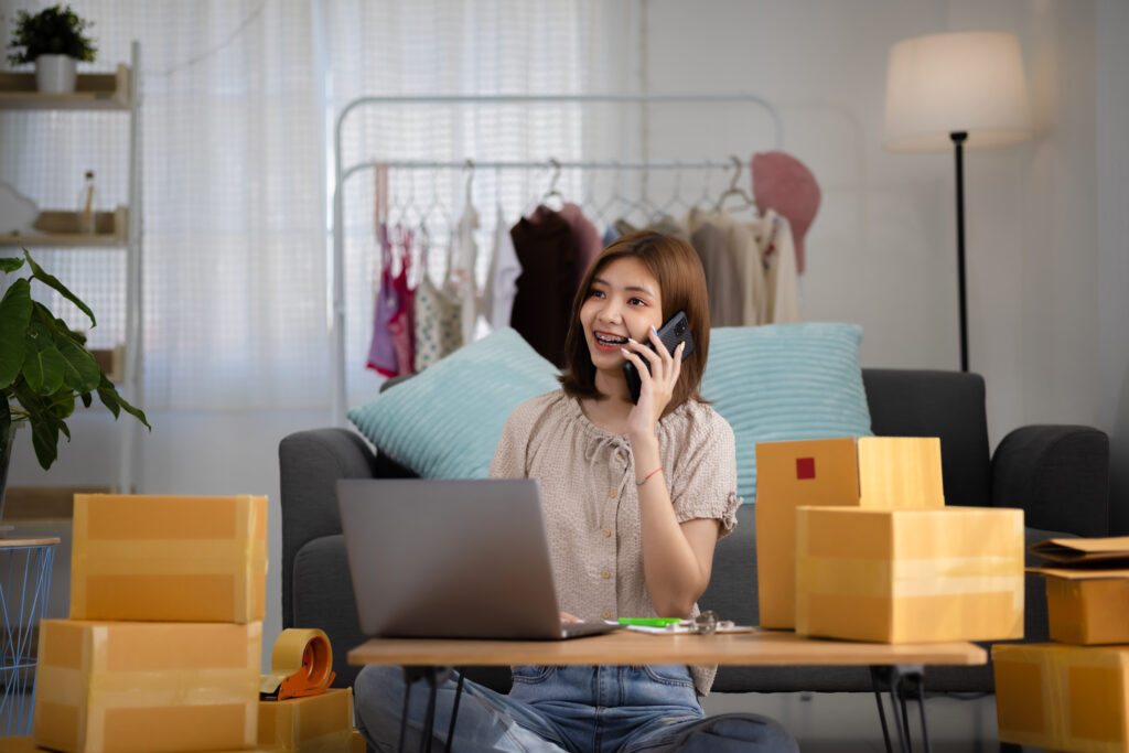 Smiling happy young Asian woman entrepreneur receiving phone call for new sales order among boxes of product with laptop computer. Success online business and eCommerce concept.